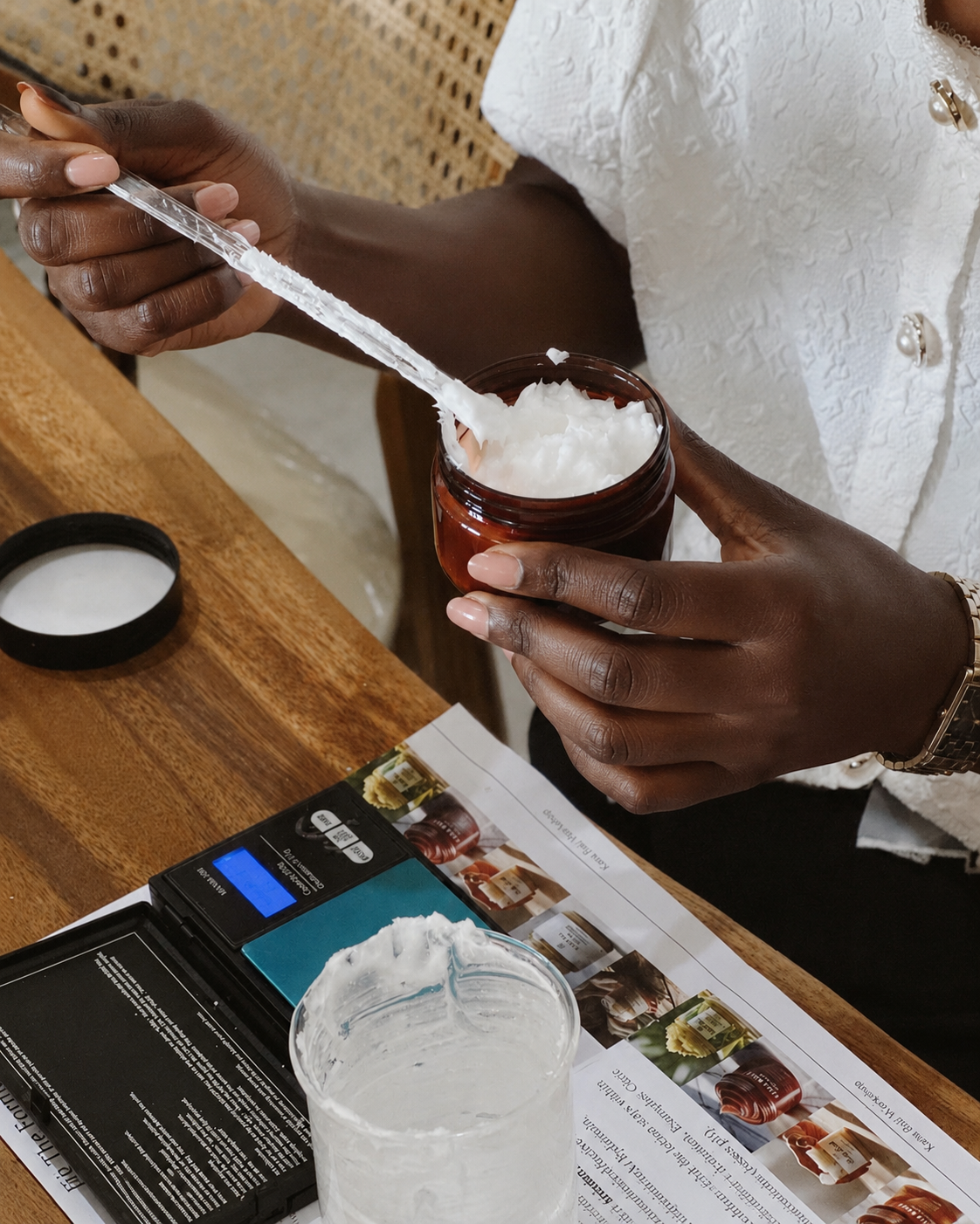 A person scoops a white cream or balm from an amber glass jar using a metal spatula, set on a wooden table with a digital scale, a clear plastic cup, and printed cards nearby.
